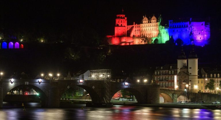 Heidelberg in bunten Lichtern bei Nacht, reflektiert im Wasser des Flusses.